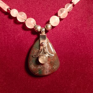 Puede incluir: Collar artesanal con un gran colgante en forma de lágrima con un engaste plateado y una piedra redonda más pequeña. El colgante cuelga de una hebra de cuentas rosas y transparentes, creando un accesorio único y elegante.
