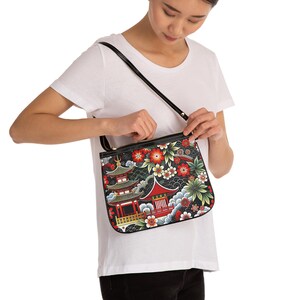 May include: A rectangular shoulder bag featuring a black background with a vibrant print of red and green pagodas, stylised flowers, and wave patterns. The bag has a black zip closure along the top and a thin black shoulder strap attached to the sides.