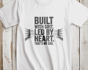 May include: White t-shirt with a crew neck and short sleeves. The shirt features the text "BUILT WITH GRIT. LED BY HEART. THAT'S MY DAD." in a bold, gray font, with a boxing ring graphic.