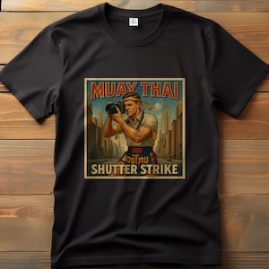 May include: Black t-shirt with a vintage-style graphic. The graphic features a man holding a camera with the text "MUAY THAI" above and "SHUTTER STRIKE" below. The image has a retro aesthetic with a cityscape background.