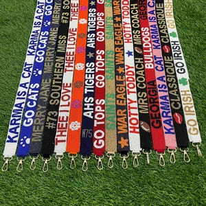 Puede incluir: Una colección de cordones con cuentas de varios colores, cada uno con texto y diseños. Los cordones tienen cierres dorados y se muestran sobre una superficie verde. El texto incluye frases como "Karma is a Cat" y "Go Irish".