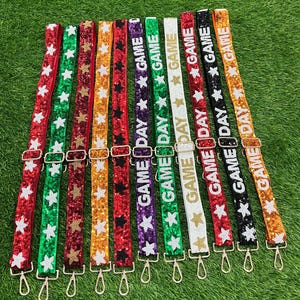 Può includere: Una collezione di tracolle per borse decorate con perline colorate con la scritta "GAME DAY" e decorazioni a stella. Le tracolle sono disponibili in vari colori, tra cui rosso, verde, oro, viola, bianco, nero e arancione, con finiture dorate.