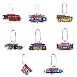 Może przedstawiać: Zestaw siedmiu breloków z logo "Scarlet & Violet" i nazwami różnych rozszerzeń. Każdy brelok ma srebrny łańcuszek i kolorowy design. Rozszerzenia obejmują "Destined Rivals", "Prismatic Evolutions" i "Surging Sparks".