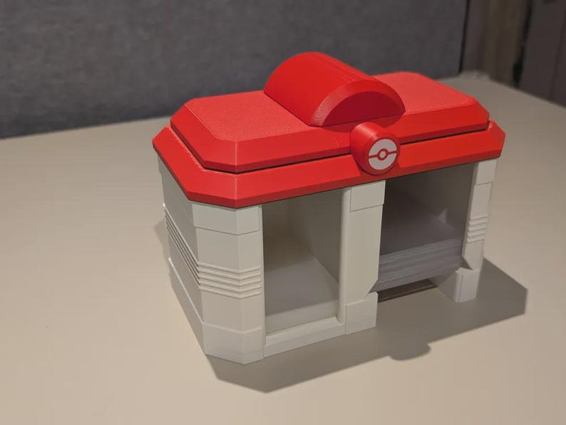 May include: A red and white storage box with a rounded top and a circular emblem. The box has a rectangular shape with open compartments. The design is reminiscent of a video game theme.