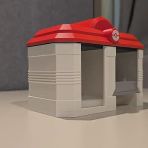 May include: A white and red 3D-printed model of a building with a red roof and a circular emblem. The building has a rectangular shape with two open sides and horizontal lines on the walls. The model is on a white surface.