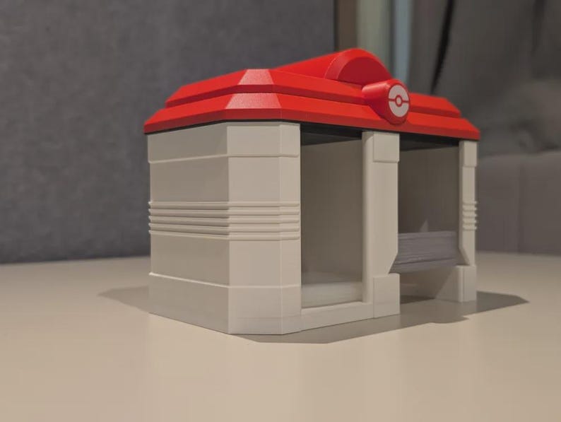 May include: A white and red 3D-printed model of a building with a red roof and a circular emblem. The building has a rectangular shape with two open sides and horizontal lines on the walls. The model is on a white surface.