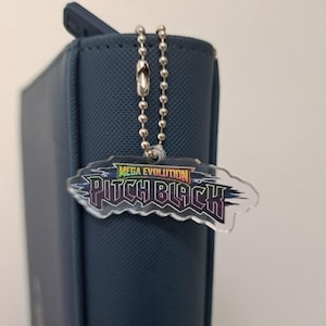 Op de afbeelding: Een doorzichtige acryl sleutelhanger met de tekst Mega Evolution Pitch Black in een gestileerd lettertype met een regenboogverloopeffect. De sleutelhanger is bevestigd aan een zilverkleurige metalen kogelketting, die om de rug van een donkerblauwe map zit.