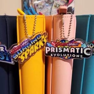 Pu&ograve; includere: Quattro raccoglitori di carte colorati, blu navy, giallo, rosa e turchese, ciascuno con un ciondolo diverso. I ciondoli riportano le scritte "Silver Tempest", "Surging Spark", "Prismatic Evolutions" e "Journey Together".