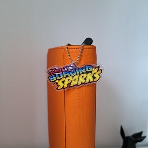 Peut inclure: Une trousse orange avec fermeture éclair, une chaîne argentée et un porte-clés en acrylique transparent. Le porte-clés affiche le texte "Scarlet & Violet Surging Sparks" en bleu, jaune et orange. La trousse est sur une surface blanche.