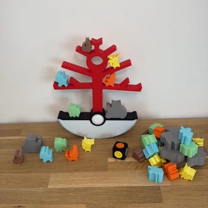 Puede incluir: Un juguete rojo en forma de árbol con figuras de animales coloridas y un dado. La base es blanca y negra. Las figuras de animales son de varios colores, incluyendo azul, verde, naranja, amarillo y gris. El juguete está sobre una superficie de madera.