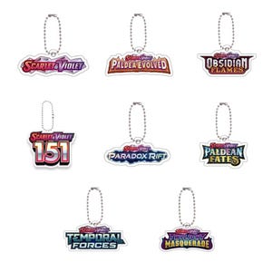 Puede incluir: Colección de siete llaveros con el logotipo "Scarlet & Violet" y varios títulos de juegos. Cada llavero tiene una cadena plateada y un dije de acrílico transparente con texto y gráficos coloridos. Los títulos incluyen "Paldea Evolved".