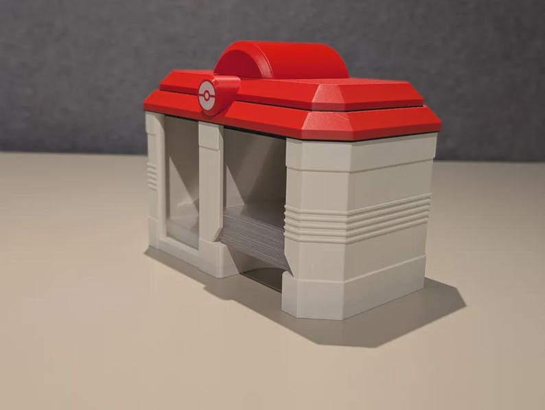 May include: A red and white, 3D-printed card holder, featuring a red roof with a white and red emblem. The card holder has a rectangular shape with a white base and a slot for cards. The design is reminiscent of a building.