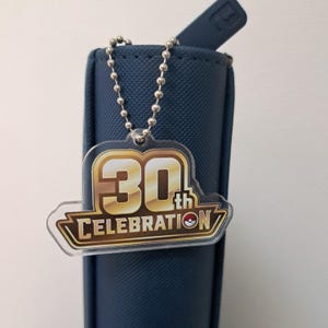 Puede incluir: Un llavero de acrílico transparente con un gráfico dorado y blanco que dice 30th CELEBRATION con un pequeño icono circular rojo y blanco. El llavero cuelga de una cadena de bolas de metal plateado sujeta a un estuche azul marino texturizado.