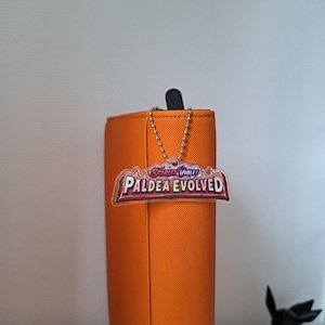 Può includere: Un raccoglitore arancione con una parte superiore nera e una catena argentata attaccata a un portachiavi trasparente Paldea Evolved. Il portachiavi ha le parole "Scarlet & Violet" e "Paldea Evolved" stampate in rosso, bianco e marrone.