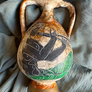 Puede incluir: Jarrón decorativo de cerámica con dos asas, con una ilustración en blanco y negro de una persona tocando la flauta. El jarrón tiene un acabado moteado marrón y blanco y una base verde y dorada.