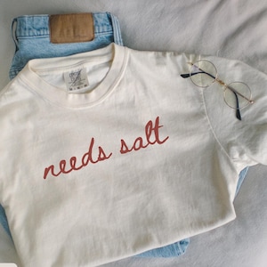 Può includere: T-shirt color avorio con la scritta "needs salt" in rosso. Abbinato a jeans blu chiaro e occhiali rotondi con montatura dorata. Gli articoli sono disposti su una superficie bianca.