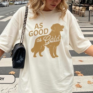 May include: Ivory-colored t-shirt featuring a golden retriever silhouette with the text "AS GOOD as Gold" in a matching gold color. The shirt has short sleeves and a relaxed fit. A black quilted shoulder bag with a chain strap is visible.