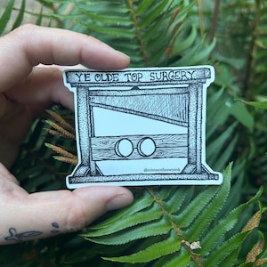 May include: A black and white sticker featuring a guillotine design with the text "YE OLDE TOP SURGERY". The sticker is held against a backdrop of green fern leaves. The sticker also includes the text "@crimsonhoneyink".
