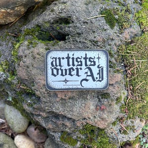 以下が含まれることがあります： ゴシック体の「artists over all」の文字が白黒の背景に配置された長方形のステッカー。ステッカーは苔で覆われた岩の上に置かれ、下部には「@crimsonhoneyink」の文字が書かれています。