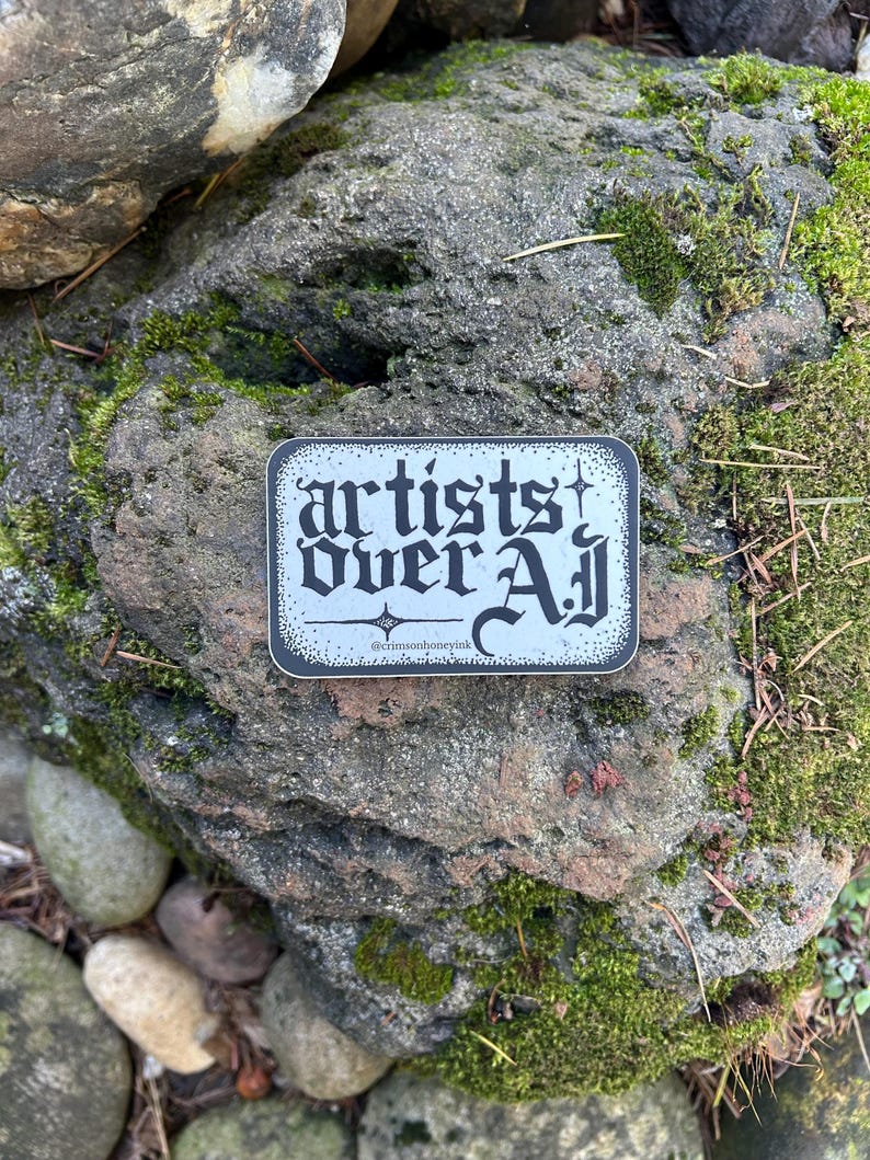 以下が含まれることがあります： ゴシック体の「artists over all」の文字が白黒の背景に配置された長方形のステッカー。ステッカーは苔で覆われた岩の上に置かれ、下部には「@crimsonhoneyink」の文字が書かれています。