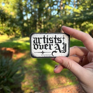 以下が含まれることがあります： ゴシック体で「artists over all」と書かれた白黒の長方形ステッカー。ステッカーには装飾的なボーダーがあり、背景はぼやけた緑色です。