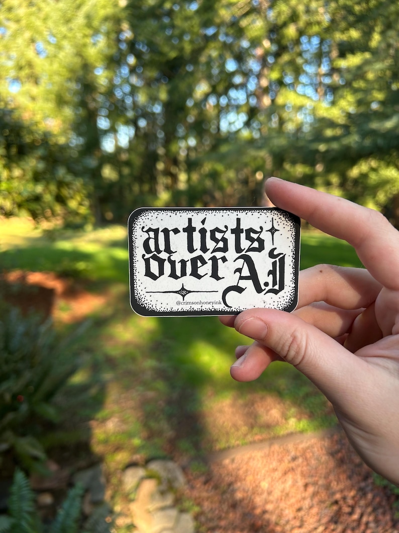 以下が含まれることがあります： ゴシック体で「artists over all」と書かれた白黒の長方形ステッカー。ステッカーには装飾的なボーダーがあり、背景はぼやけた緑色です。