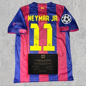Puede incluir: Una camiseta de fútbol del Barcelona con rayas verticales azules y rojas. Muestra "NEYMAR JR" y el número "11" en amarillo. Incluye un parche "Respect" y un logo de la Champions League. Se muestra un certificado de homenaje.