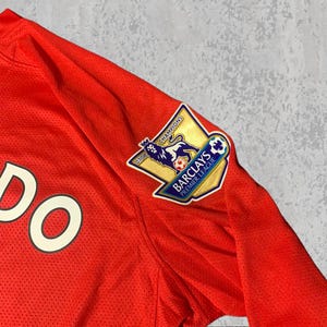 Pu&ograve; includere: Primo piano di una maglia da calcio rossa con la parola "DO" visibile. La maglia presenta una toppa della Premier League Barclays con un emblema di leone e la scritta "Champions 07/08". Il tessuto &egrave; traspirante e strutturato.