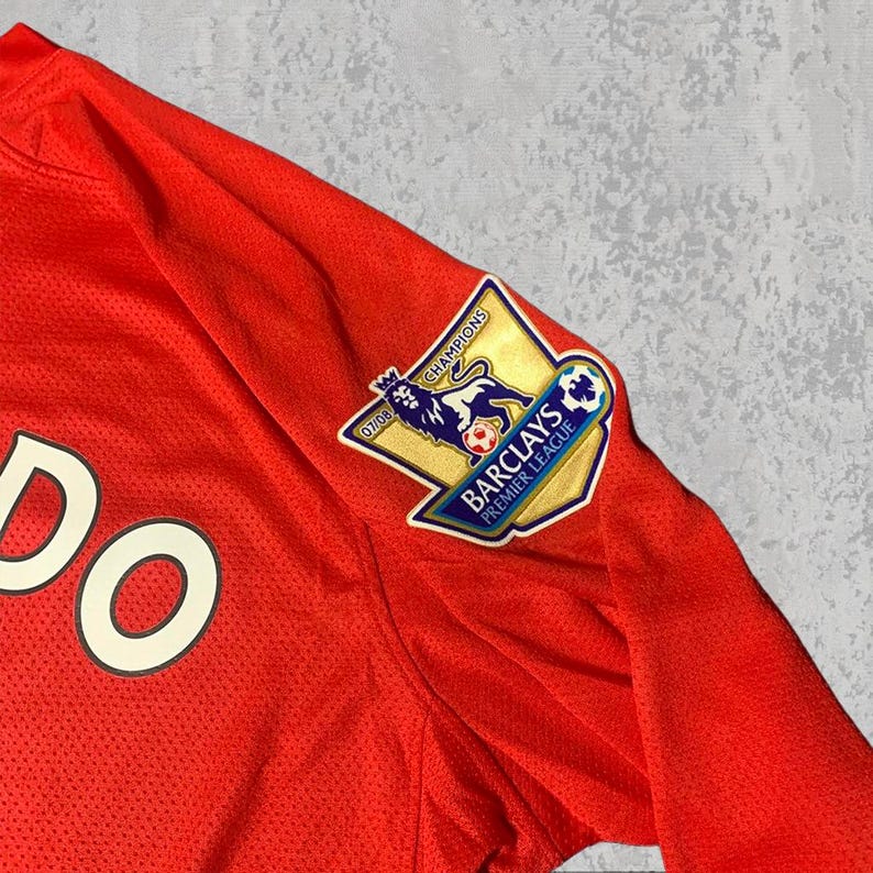 Pu&ograve; includere: Primo piano di una maglia da calcio rossa con la parola "DO" visibile. La maglia presenta una toppa della Premier League Barclays con un emblema di leone e la scritta "Champions 07/08". Il tessuto &egrave; traspirante e strutturato.
