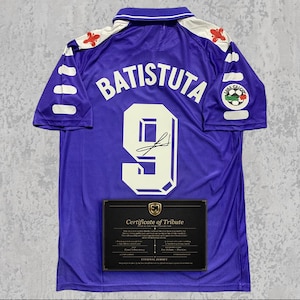 May include: A purple football shirt with the name "BATISTUTA" and the number "9" on the back. The shirt has white detailing on the shoulders and sleeves, and a certificate of tribute is displayed below. The shirt also has a team logo on the right sleeve.