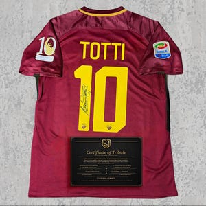 Puede incluir: Camiseta de fútbol granate con el nombre "TOTTI" y el número "10" en amarillo. La camiseta tiene cuello y detalles de manga dorados, con un parche "SERIE A TIM". Un certificado de homenaje se muestra debajo.