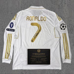 Può includere: Maglia da calcio bianca a maniche lunghe con dettagli dorati, il nome "RONALDO" e il numero "7" in oro. Include un "Certificato di Tributo" e una toppa della Champions League UEFA.