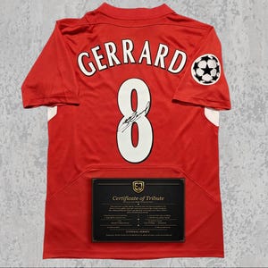 Può includere: Maglia da calcio rossa con il nome "GERRARD" e il numero "8" in bianco. Presenta una toppa della Champions League e un certificato di tributo. Una maglia commemorativa.