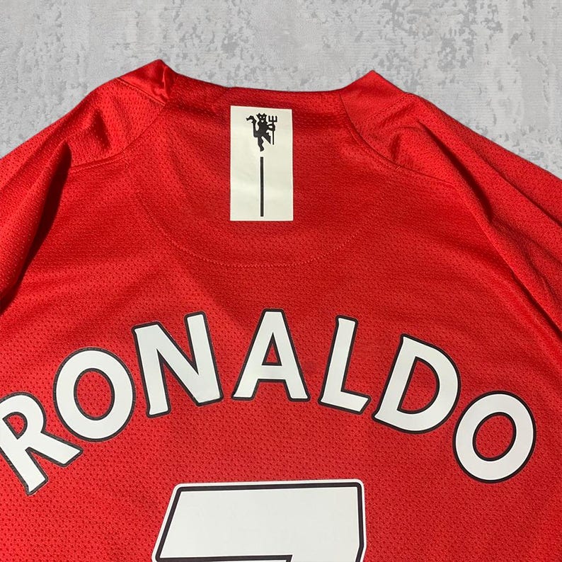 Pu&ograve; includere: Maglia da calcio rossa con il nome "RONALDO" a lettere bianche sul davanti. La maglia presenta una toppa rettangolare bianca con un emblema nero e una linea nera verticale. Tessuto a rete traspirante.