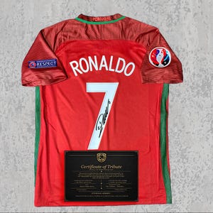 May include: A red Portugal football shirt with "RONALDO" and the number "7" in white. The shirt has green side stripes and a "RESPECT" patch on the left sleeve. A certificate of tribute is displayed below the jersey.