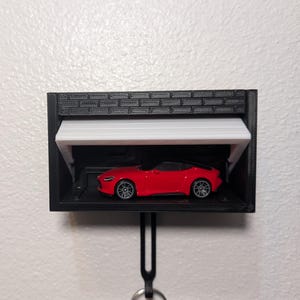 May include: A miniature red sports car is displayed inside a black garage-style key holder. The garage has a white, hinged door and a brick-patterned top. The key holder is hanging on a white wall.