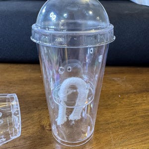 May include: A clear plastic cup with a domed lid. Inside, a white, U-shaped object is suspended. The cup has a ridged rim and small holes. The cup is sitting on a wooden surface.