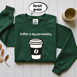 Könnte beinhalten: Waldgrünes Sweatshirt mit dem Text "Coffee is my personality" und einer Kaffeetassen-Grafik. Das Sweatshirt hat eine einfarbige waldgrüne Farbe. Auf der Vorderseite ist eine Kaffeetasse mit einer schwarzen Tasse und einer weißen Kaffeebohne aufgedruckt.
