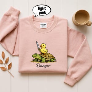 May include: Light pink Gildan sweatshirt featuring a cartoon duckling riding a turtle and holding a sword. The word "Danger" is below the graphic. A speech bubble above the sweatshirt says "light pink".