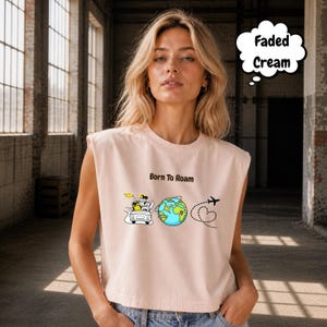 May include: Faded cream-colored sleeveless crop top with the text "Born To Roam" and travel-themed graphics. The graphics include a car, a globe, and an airplane with a heart-shaped flight path.