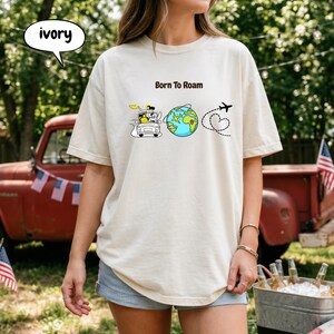 May include: Ivory-colored t-shirt with the text "Born To Roam" and a graphic of a car, globe, airplane, and heart. The shirt is a relaxed fit and suitable for casual wear.
