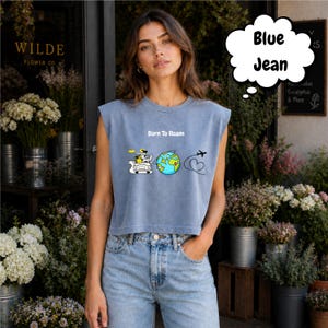 May include: A light blue crop top with the text "Born To Roam" and a graphic of a car, globe, and airplane. The top is paired with blue jeans. The image is taken outside a flower shop.