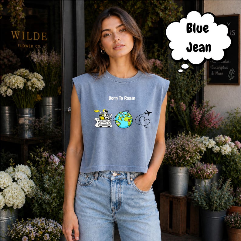 May include: A light blue crop top with the text "Born To Roam" and a graphic of a car, globe, and airplane. The top is paired with blue jeans. The image is taken outside a flower shop.