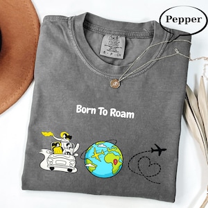 May include: A gray Comfort Colors t-shirt with the words "Born To Roam" in white. The graphic includes a car with passengers, a globe with a plane, and a dotted line with a heart and airplane. A silver necklace with a pendant is on the shirt.