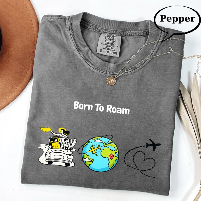 May include: A gray Comfort Colors t-shirt with the words "Born To Roam" in white. The graphic includes a car with passengers, a globe with a plane, and a dotted line with a heart and airplane. A silver necklace with a pendant is on the shirt.