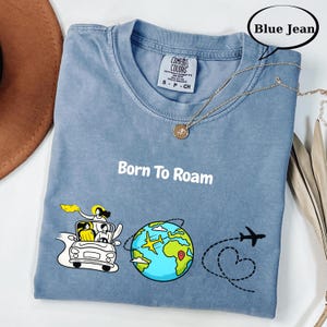 May include: Blue jean colored t-shirt with the text "Born To Roam" and a graphic of a car, globe, and airplane. The shirt has a crew neck and short sleeves. A gold necklace is draped across the shirt.
