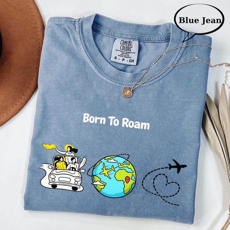 May include: Blue jean colored t-shirt with the text "Born To Roam" and a graphic of a car, globe, and airplane. The shirt has a crew neck and short sleeves. A gold necklace is draped across the shirt.