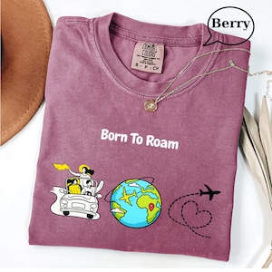 May include: Berry-colored t-shirt with the text "Born To Roam" and a graphic of a car, globe, and airplane. The shirt has a crew neck and a small tag. A silver necklace with a pendant is draped across the shirt.