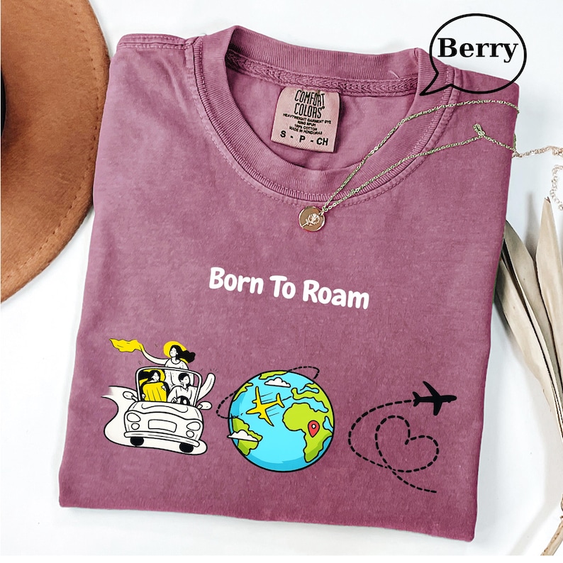 May include: Berry-colored t-shirt with the text "Born To Roam" and a graphic of a car, globe, and airplane. The shirt has a crew neck and a small tag. A silver necklace with a pendant is draped across the shirt.