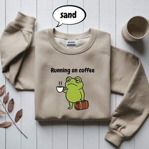 May include: Sand-colored sweatshirt with the text "Running on coffee" and a cartoon frog holding a coffee cup and suitcase. The frog is green with a tired expression. A neutral-toned mug is in the background.